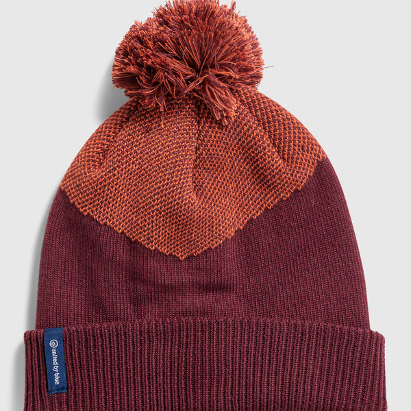 Eggplant Recycled Novelty Pom Beanie (Flat-lay): Deep burgundy knit beanie with orange textured chevron panel and pom-pom top