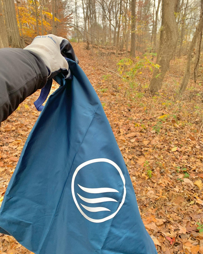 Navy Reusable DIY Cleanup Kit (Lifestyle): Gloved hand holds folded navy bag with white wave logo in autumn forest