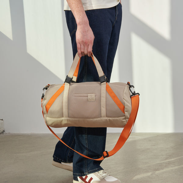 Sand (Re)active™ 16L Mini Duffle (In-use): Person holding tan duffle with orange straps and carabiner clips at side