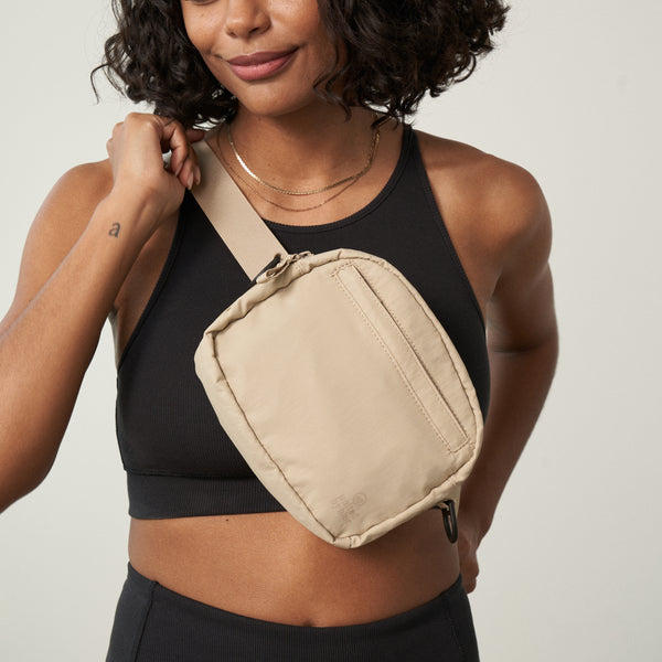 Sand (Re)active™ 2L Convertible Crossbody (In-use): Woman wearing beige crossbody sling bag diagonally over black athletic top