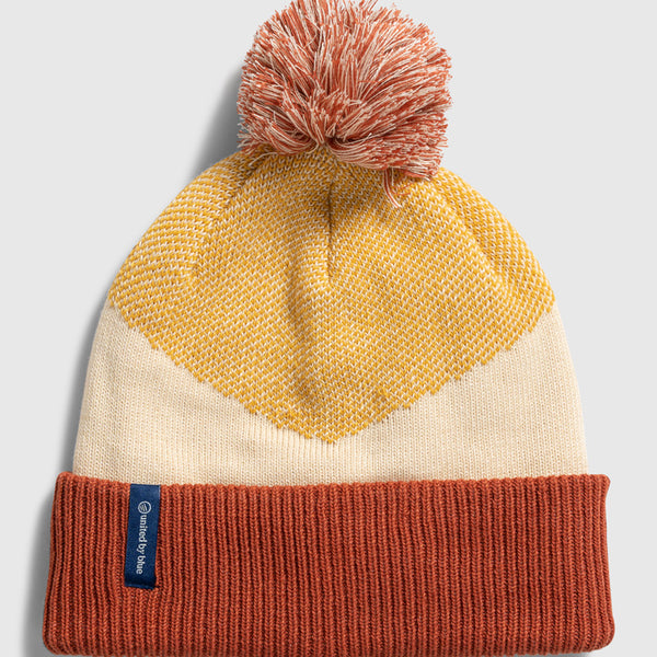 DANDELION Recycled Chevron Pom Beanie (Flat-lay): Rust cuff beanie with cream body, gold chevron pattern, and rust pom-pom