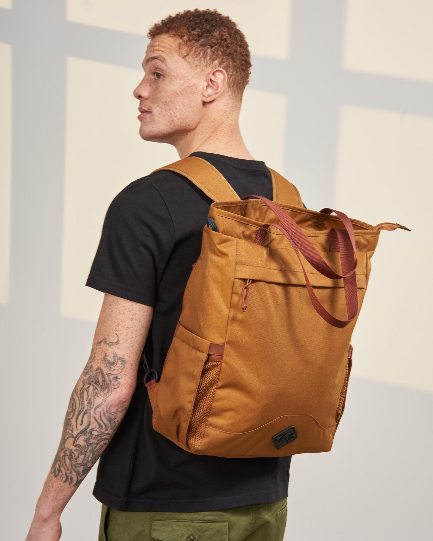 Camel (R)evolution™ 25L Convertible Carryall (In-use): Man wearing tan convertible backpack with rust straps, side mesh pocket visible