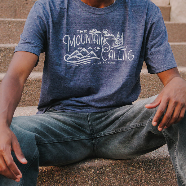 Heathered Navy Mountains Are Calling - Biodegradable Graphic Tee - (All-gender) (In-use): Man sitting on stone steps wearing navy graphic tee with mountain print