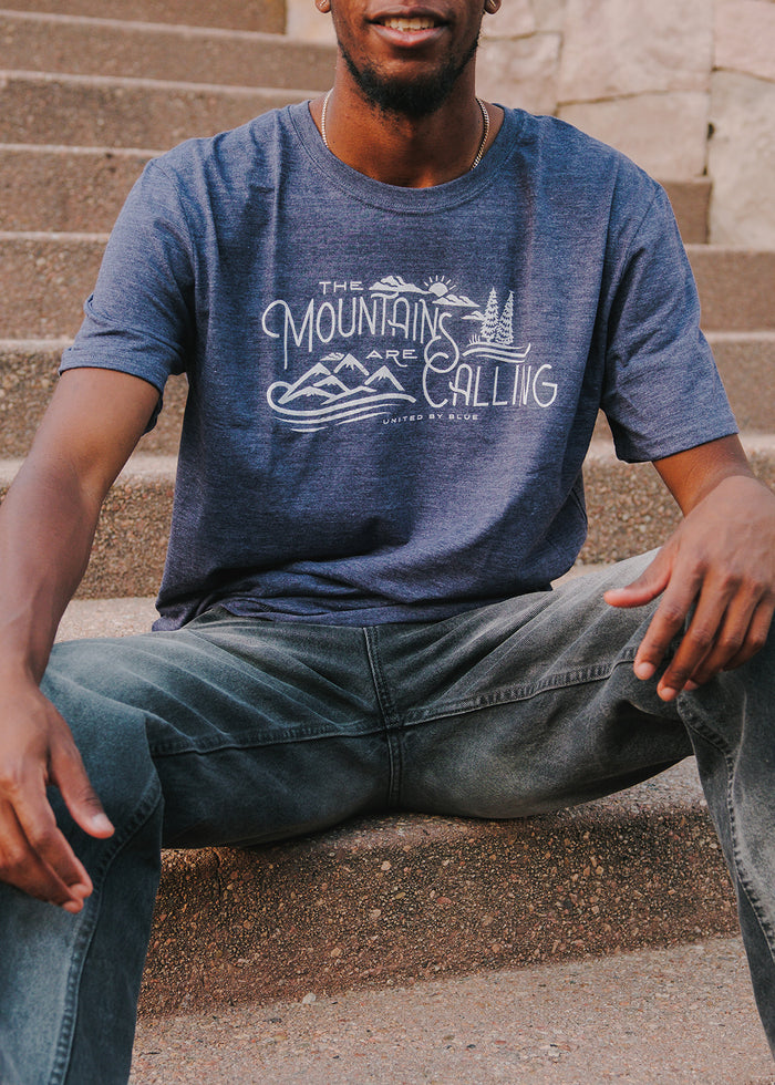 Heathered Navy Mountains Are Calling - Biodegradable Graphic Tee - (All-gender) (In-use): Man sitting on stone steps wearing navy graphic tee with mountain print