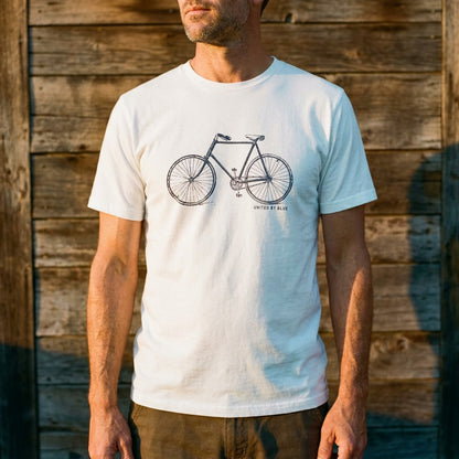 Antique White Unisex Graphic Tee in 100% Organic Cotton - Vintage Bicycle (Lifestyle): Man in cream bicycle-graphic tee against a rustic wooden plank wall