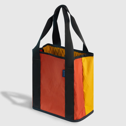 Coral Red The Westward Tote (Angle): Orange and yellow tote with black straps, trim, and blue label