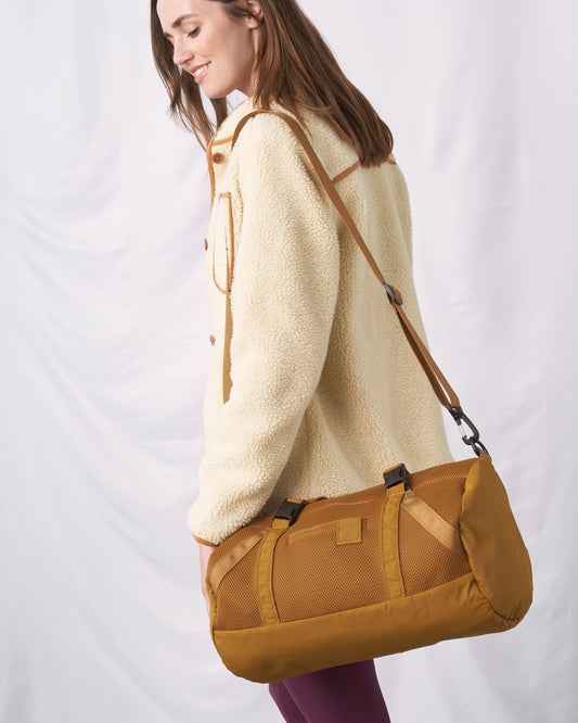 Smoky Orchre (Re)active™ 16L Mini Duffle (In-use): Woman in sherpa fleece carries golden-tan duffle with crossbody strap