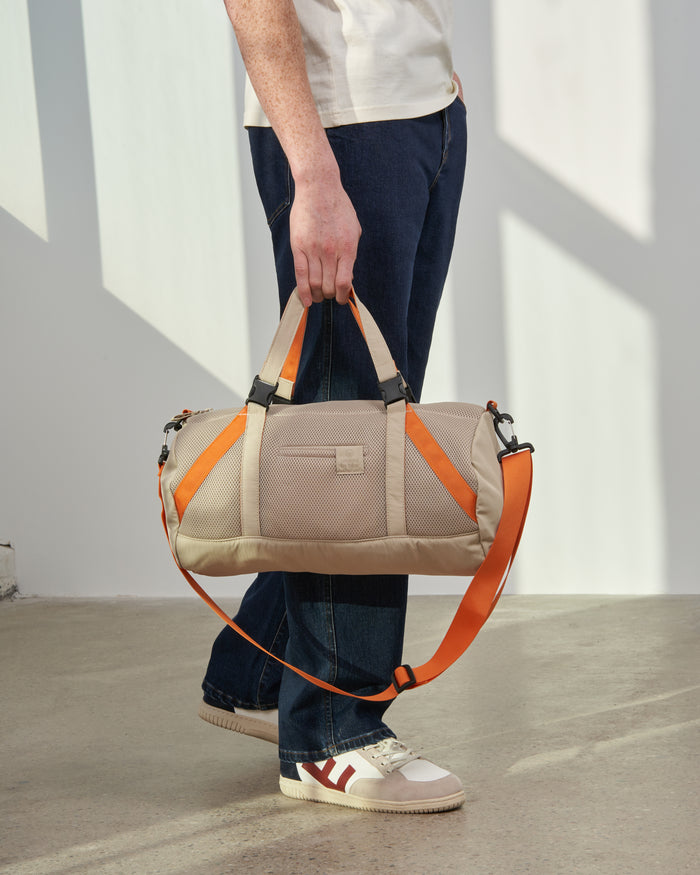 Sand (Re)active™ 16L Mini Duffle (In-use): Person holding tan duffle with orange straps and carabiner clips at side