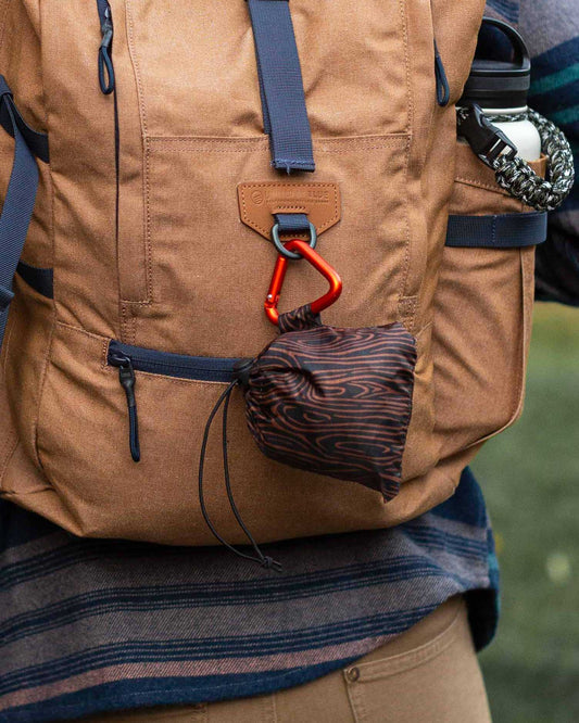 Waves The Packable Tote (In-use): Dark wave-patterned stuff sack clipped via red carabiner to tan backpack