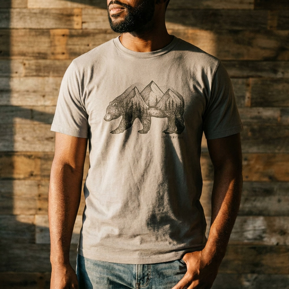 Antique White Unisex Graphic Tee in 100% Organic Cotton - Grizzly (Lifestyle): Man in cream grizzly-mountain graphic tee against a sunlit rustic wood wall