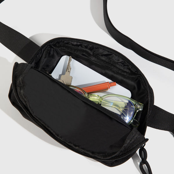 Black (Re)volution™ 1L Go-Anywhere Bag (Detail): Black fanny pack open, showing glasses, keys, phone, and orange pen inside