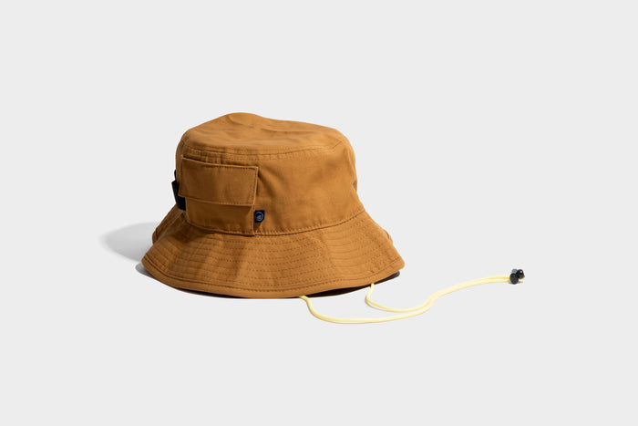 HONEY UTILITY BUCKET HAT (Angle): Tan canvas bucket hat with side pocket, chin cord, and logo patch