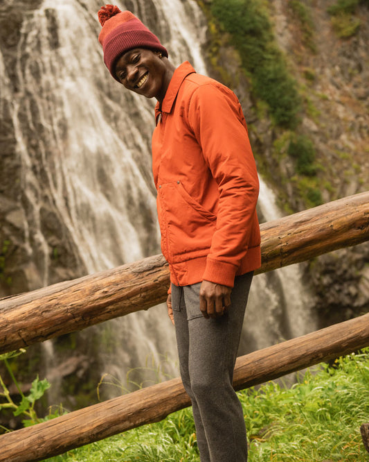 Recycled Chevron Pom Beanie (In-use): Person wearing red pom beanie leaning on wooden fence near waterfall