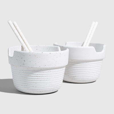 Eggshell Stoneware Stackable Noodle Bowl Set (Angle): Two speckled white ribbed bowls with matching chopsticks resting inside notched rims