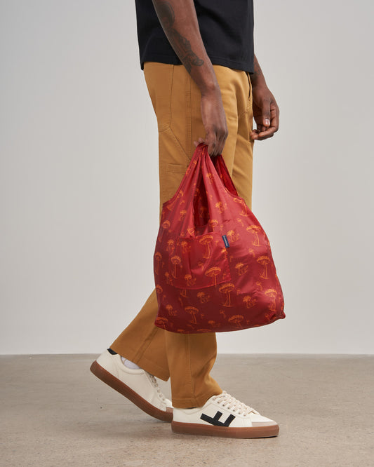 The Packable Tote (In-use): Red reusable tote with orange mushroom print, held by walking person